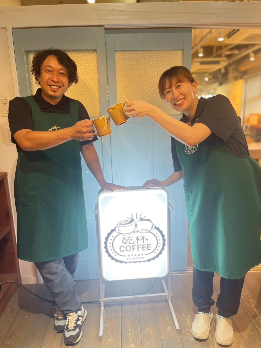 乾杯COFFEEの看板の横で、マグカップで乾杯するエプロン姿のスタッフ2人