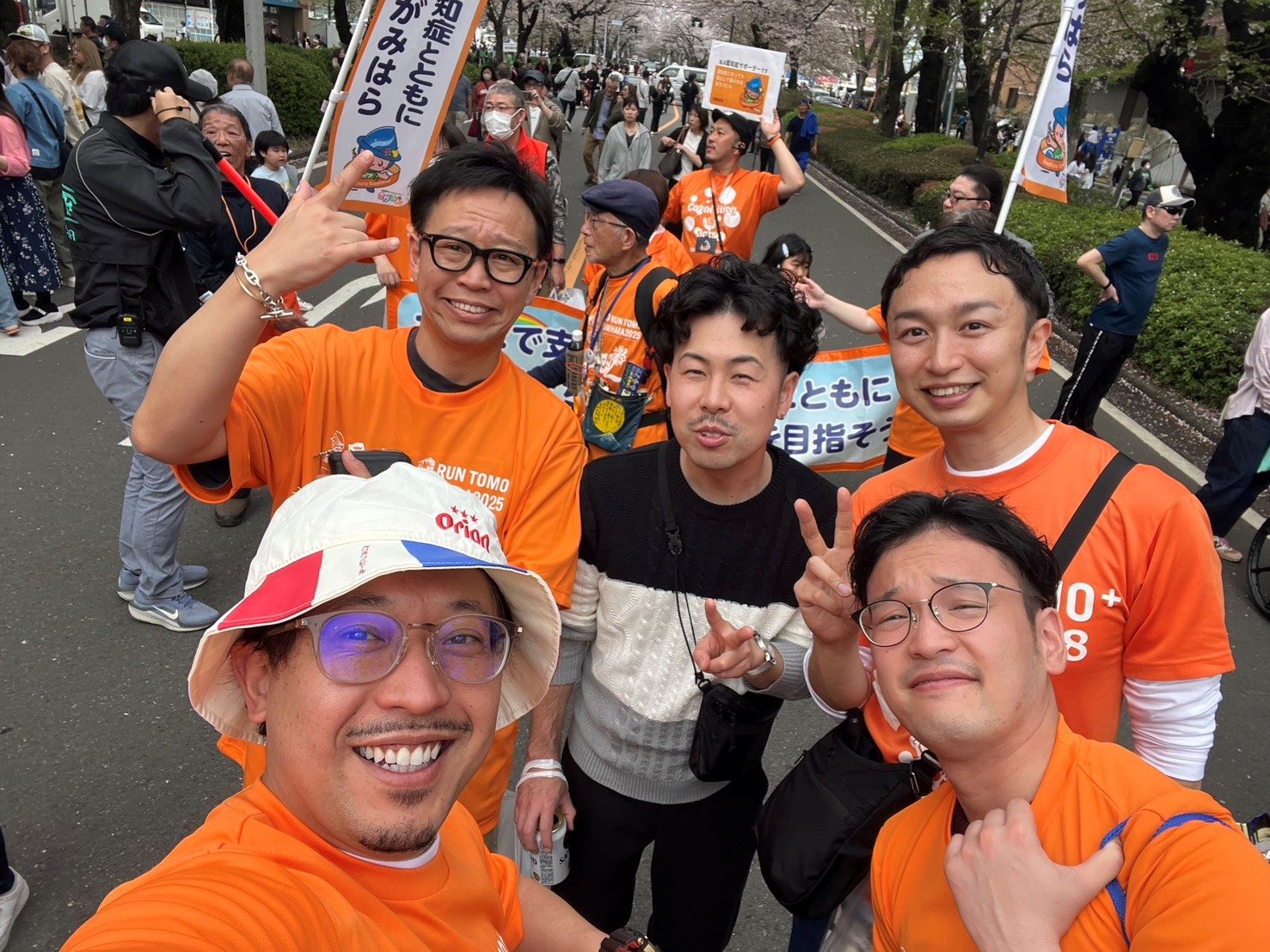 桜の下、オレンジのTシャツで笑顔の自撮りをする仲間たち